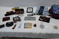 Wide shot of all lot items laid out on white cloth including sunglasses, wallets, scarves, binoculars, cosmetic bag, key rings, compact mirrors, art piece, luggage tag, and small accessories.