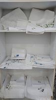 Six sets of fine linen linens with hand embroidery displayed folded on shelves, white with floral motifs in green and blue