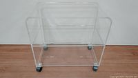 Front and side view of clear lucite cart showing minimalist open rectangular frame and caster wheels.