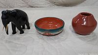Three items displayed: black hand-carved elephant sculpture with ivory tusks, round glazed pottery bowl with turquoise and rust geometric design, and red rounded pottery pot with narrow neck