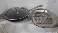 Calphalon frying pan with handle and glass lid next to two Linkfair stainless steel BBQ baskets with handles and perforations.