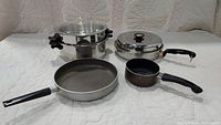 Photo showing all four cookware items: stainless steel double boiler with glass lid, large fry pan, smaller fry pan with lid, and small saucepan, all set on white quilted background.