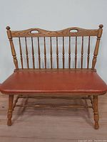 Front view of wooden bench showing turned spindle backrest, carved top rail with cutout handles, and brown leather seat cushion.
