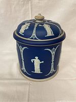 Front view of blue stoneware jar and lid with white classical reliefs on blue background and central figure shown.