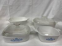 Four white Corningware baking dishes, two with clear glass lids, arranged on a white backdrop showing the blue floral design on the side.