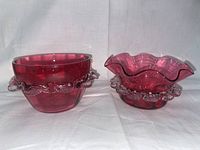 Photo showing two vintage Victorian cranberry glass dishes side by side on white cloth background. The left dish is round with a clear ruffled edge and the right dish has a wavy rim with clear scalloped glass decoration.