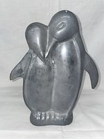 Front view of metal penguin sculpture showing the two penguins closely embraced, details of flippers and webbed feet visible. Matte metallic finish with patina and surface wear.