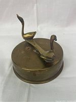 Front angled view of the round brass music box with two swan figurines mounted on top, showing patina and wear on the brass surface.