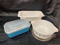 Three vintage Pyrex dishes shown together: a white and blue rectangular loaf pan, a blue rectangular casserole with a ribbed glass lid, and a round white casserole with blue garland snowflake pattern and a clear glass lid.