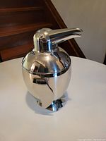 Closed penguin-shaped metal ice bucket with shiny silver surface standing on table near stairs.