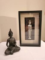 Two-item lot including black resin seated Buddha in a meditative pose holding a bowl alongside a wooden frame shadow box art featuring a silver-toned standing Buddha figurine.