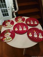 All six Christmas platers on a round table showing three large and three medium platers with festive snowman and red background designs.