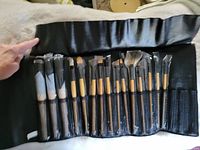 Photo of 15 makeup brushes with wooden handles in a black leather-like foldable case, each brush individually wrapped in plastic.