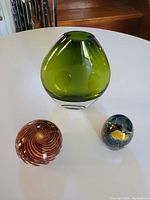 Green glass rounded vase with polished top, placed on white surface with two smaller glass objects.