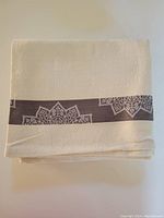 Folded off white cotton jacquard tablecloth showing subtle woven pattern and taupe border with floral designs