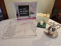 Four items arranged on a table: etched glass serving platter with leaf etching, black felt letter board sign in box, case of vanilla cream votive candles on a green holder, and vintage silver syrup pitcher with attached drip tray.
