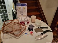 Photo of all items for sale on a round white table including cheese grater, candle, ramekin, dish cloths, battery lights, and wire racks