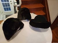 Three vintage hats on a white round table near wooden stairs: a felt hat with mink trim and two full mink hats.