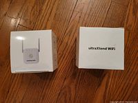 Front and top views of two unopened UltraXtend Wifi Booster boxes on wooden floor, showing device image and branding.