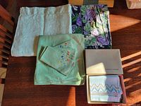 All linens spread and folded on table: two vintage embroidered tea towels in original Eaton's box, pale green damask tablecloth, purple/green/black floral cotton tablecloth, green embroidered tablecloth with matching napkins folded