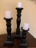Three black wooden pillar candlesticks with white pillar candles placed on a wooden surface against a light wall background, showing their relative sizes and turned design detail.