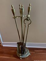 Full view of four brass fireplace tools with turned handles and finials in a round brass stand on wooden floor.