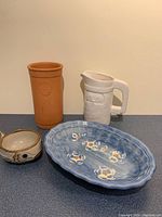 Four ceramic items shown: brown cylindrical clock, blue oval plate, white pitcher, fish-shaped cup on a gray surface with beige background.