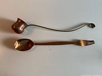 Copper spoon-fork hybrid utensil with bent fork tines and copper brandy warmer with long handle and brass 'BRANDY' label, both laid flat against a white background