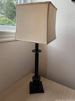 Full view of tall black table lamp with beige square shade near window.