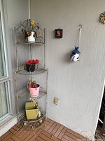Metal outdoor shelving rack holding four ceramic planters, positioned in a corner next to window and wall, one hanging ceramic bird feeder visible.