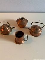 Four miniature copper household items placed on a flat surface: two kettles, one lidded pot, one tankard mug