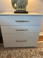 Front view of one white bedside table showing three drawers with horizontal rectangular metal handles.