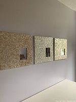 Three square wall mirrors with varied mother of pearl tile frames hung side by side on a neutral colored wall. Each has a small inset rectangular mirror at its center.