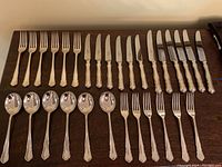 Full view of the flatware pieces laid out: six dinner forks, six dinner knives, six soup spoons, six luncheon forks, and six luncheon knives on a table surface.