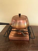 Full view of copper fondue pot with handle on copper burner base and black metal rack.