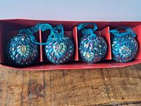 Four round blue Christmas ornaments decorated with multi-colored jewels in a red box.