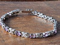 Amethyst tennis bracelet laid flat showing 12 emerald cut purple stones and decorative silver links against wooden background to highlight detail and clasp closure.
