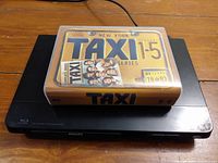 Philips Blu-Ray DVD player with Taxi TV series DVD box set on top, showing box set cover and player surface.