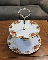 Photo of a two tiered cake tray showing white porcelain plates with floral pink and yellow flower patterns around the edges and gold trim. Silver metal handle in the center connects the plates and provides carrying functionality.