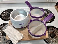 Wilton electric candy melting pot base unit with two purple handled double-sided sieves and multiple white lollipop sticks on a wooden board.
