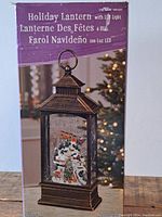 Boxed lantern showing holiday scene with Santa, sleigh, reindeer, and Christmas tree inside a bronze-colored vintage lantern with LED light feature.