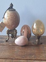 Four decorative eggs on wooden surface including large Bombay stone egg on brass elephant leg stand, carved pink egg from Kenya, polished pinkish-brown egg, and translucent yellow egg on brass stand.