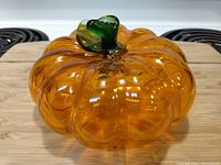 Orange glass pumpkin figurine with a green stem on a wooden surface, stove burners in background