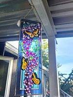 Vertical stained glass panel hanging outdoors showing yellow butterflies and purple flowers with blue background.