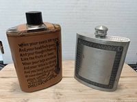 Pair of flasks on wooden surface showing full view of both items side by side; glass flask has tan leather cover with printed wording and illustration, metal flask is plain with etched border.