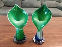 Pair of uniquely shaped glass vases with green interior and gradient exteriors from green to black and green to blue. Positioned side by side on a wooden surface with a couch in the background.