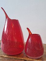 Two red glass vessels on wooden surface, larger and smaller with pointed spouts