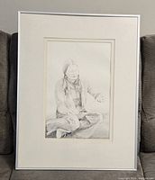 Framed pencil drawing showing a woman sitting cross-legged, holding an object, with fine shading and detail.