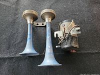 Front view of two blue metal trumpet horns with grey plastic caps and black cylindrical compressor attached separately on dark background.