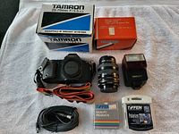 Overview photo showing Canon T70 camera body, Tamron 28-70mm lens, Kitstar zoom lens, Sunpak 277T flash, camera cable, two lens filter boxes and Tiffen polarizer filter case arranged on towel.